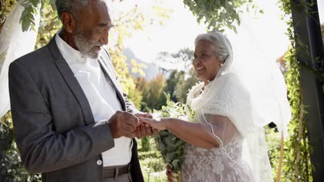 happy senior biracial couple exchanges rings during wedding ceremony in garden, slow motion