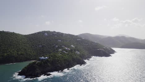 Cautivadora-Vista-Aérea-Rodeando-La-Isla-Virgen-Usvi-Playa-De-Arena-Blanca-Cielo-De-Agua-Azul-Nubes-Blancas-Agua-Turquesa-Con-Casas-En-El-Acantilado-De-La-Montaña