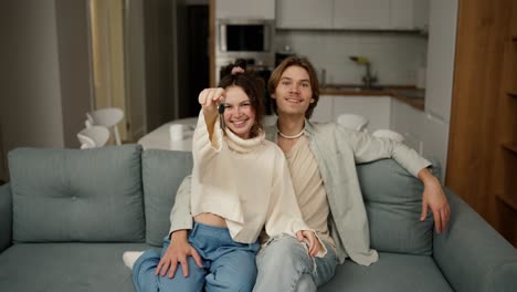 smiling millennial couple home owners sitting on cozy sofa, showing keys to camera, feeling excited