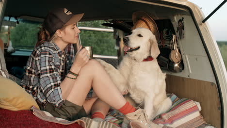 young woman drinking tea and petting golden retriever in van