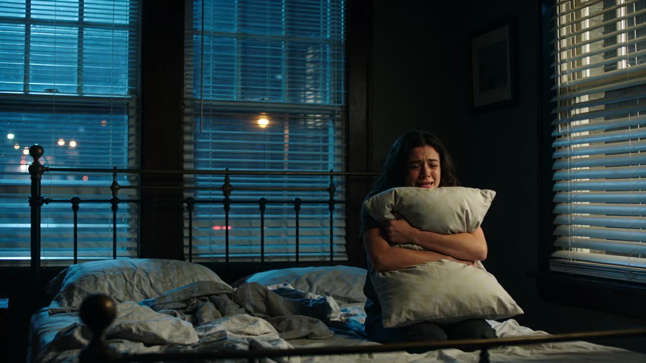 Woman starting hugging pillow on bed, city lights shining through windows, crying, seeking health