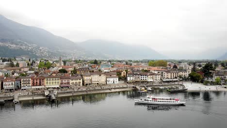 Sobrevuelo-Aéreo-A-Lo-Largo-Del-Paseo-Marítimo-De-Ascona-En-Ticino,-Suiza,-Con-Vistas-A-Un-Barco-Que-Llega-Al-Muelle-Y-A-Los-Edificios-De-La-Ciudad-A-Lo-Largo-Del-Paseo-Junto-Al-Lago-A-Orillas-Del-Lago-Maggiore