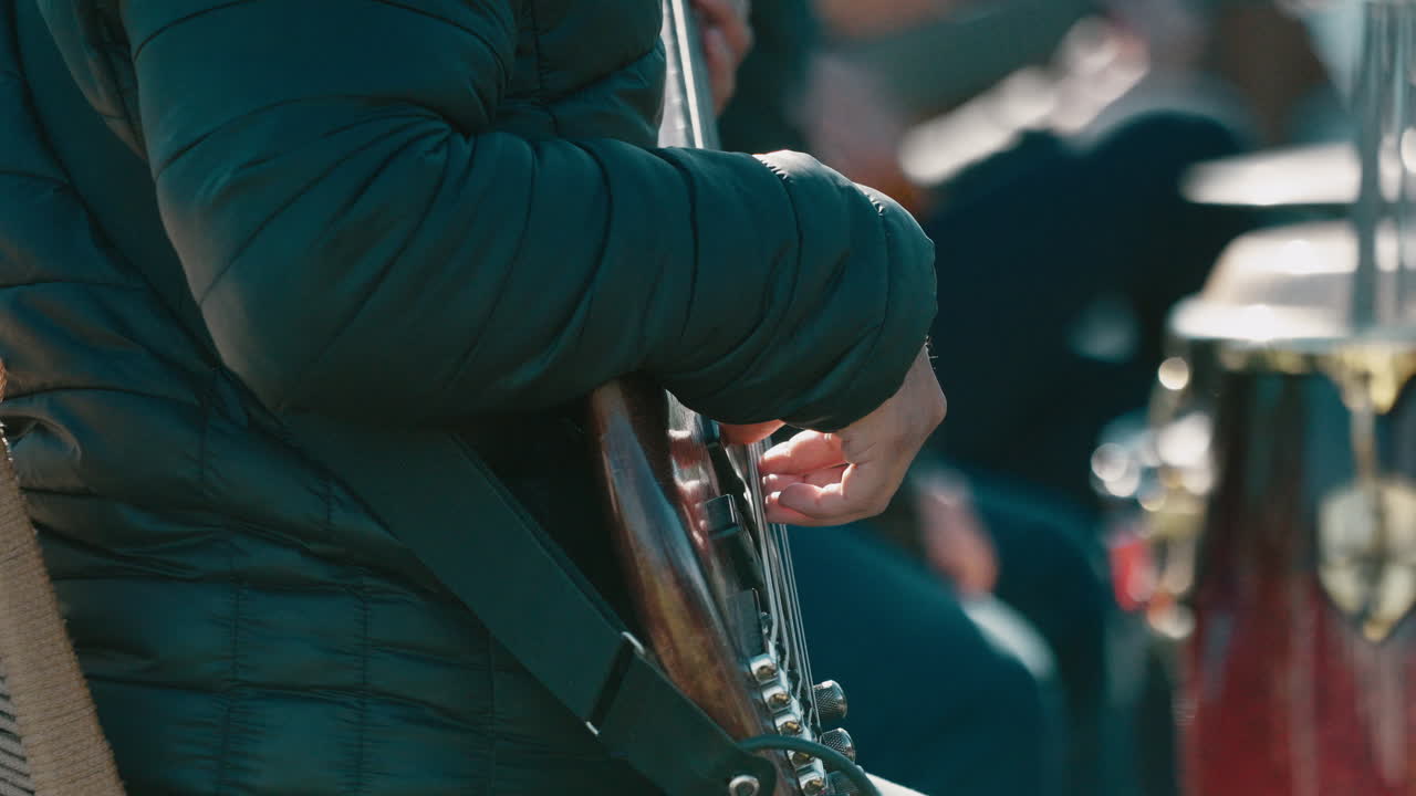 Musicista che indossa una giacca imbottita suona un basso elettrico all'aperto, primo piano si concentra sulla mano destra che pizzica le corde e sul corpo in legno lucido, luce naturale, tempo reale, inquadratura statica