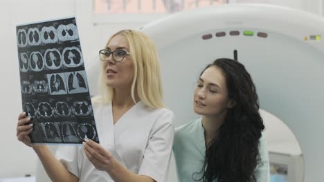 woman doctor radiologist explains the results of ct scanning for young female patient, showing the snapshot with images, observing and analyzing ct scan in modern clinic beside modern ct scanner