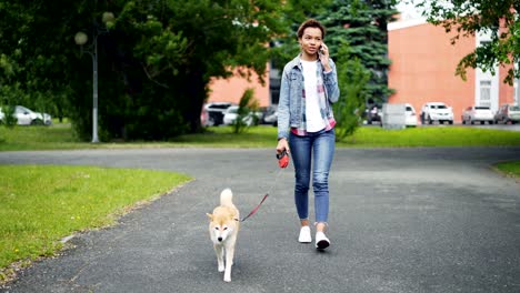 happy mixed race girl is walking dog in park and talking on mobile phone communicating with friends. modern technology, loving animals and communication concept.