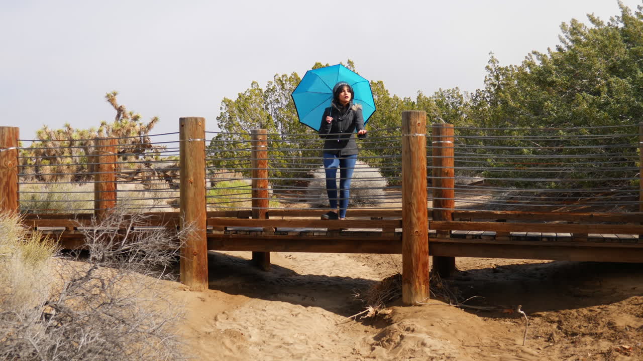 una mujer bonita con un paraguas azul parado en un puente en una reserva natural del desierto durante una tormenta de lluvia ligera a cámara lenta
