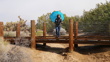 una mujer bonita con un paraguas azul parado en un puente en una reserva natural del desierto durante una tormenta de lluvia ligera a cámara lenta