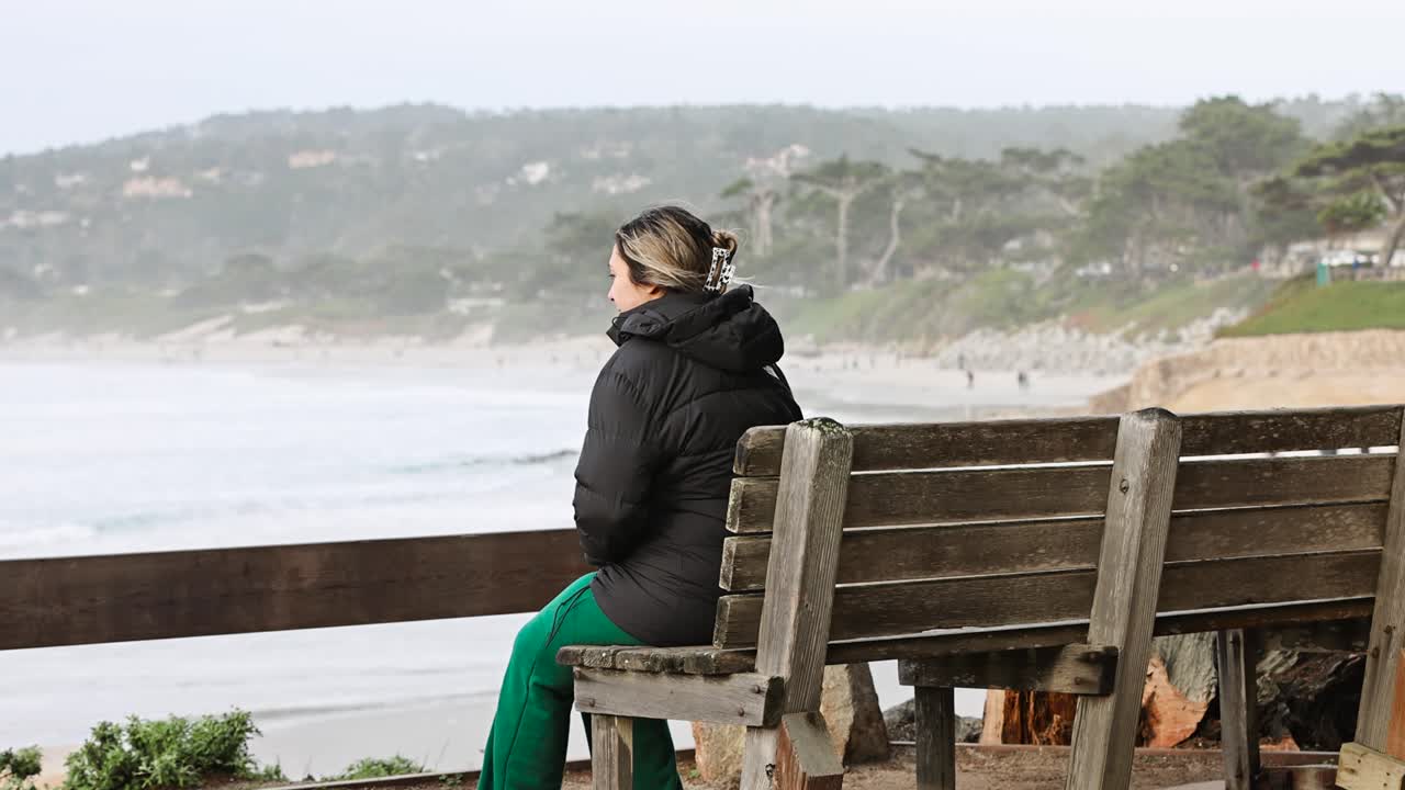 young girl in a black down jacket sitting on a bench taking in Carmel by the sea beach seagull flyby STATIC SHOT