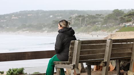 young girl in a black down jacket sitting on a bench taking in carmel by the sea beach seagull flyby static shot