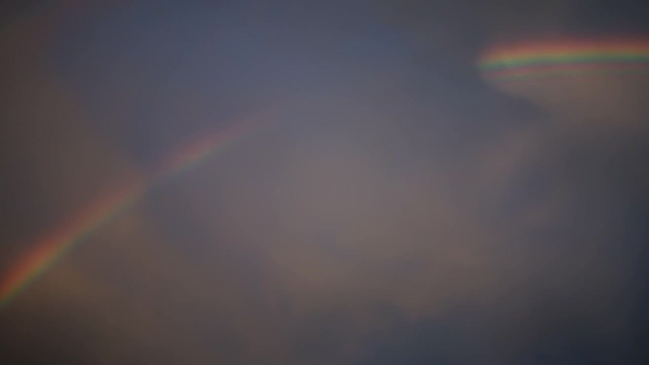 Dynamic handheld footage captures a vivid rainbow arching across dramatic, cloudy skies in the USA. Soft natural light and fluid camera motion evoke a serene, magical atmosphere.