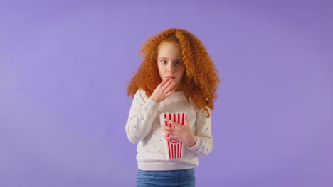 scared girl watching horror movie and spilling popcorn at cinema against purple background