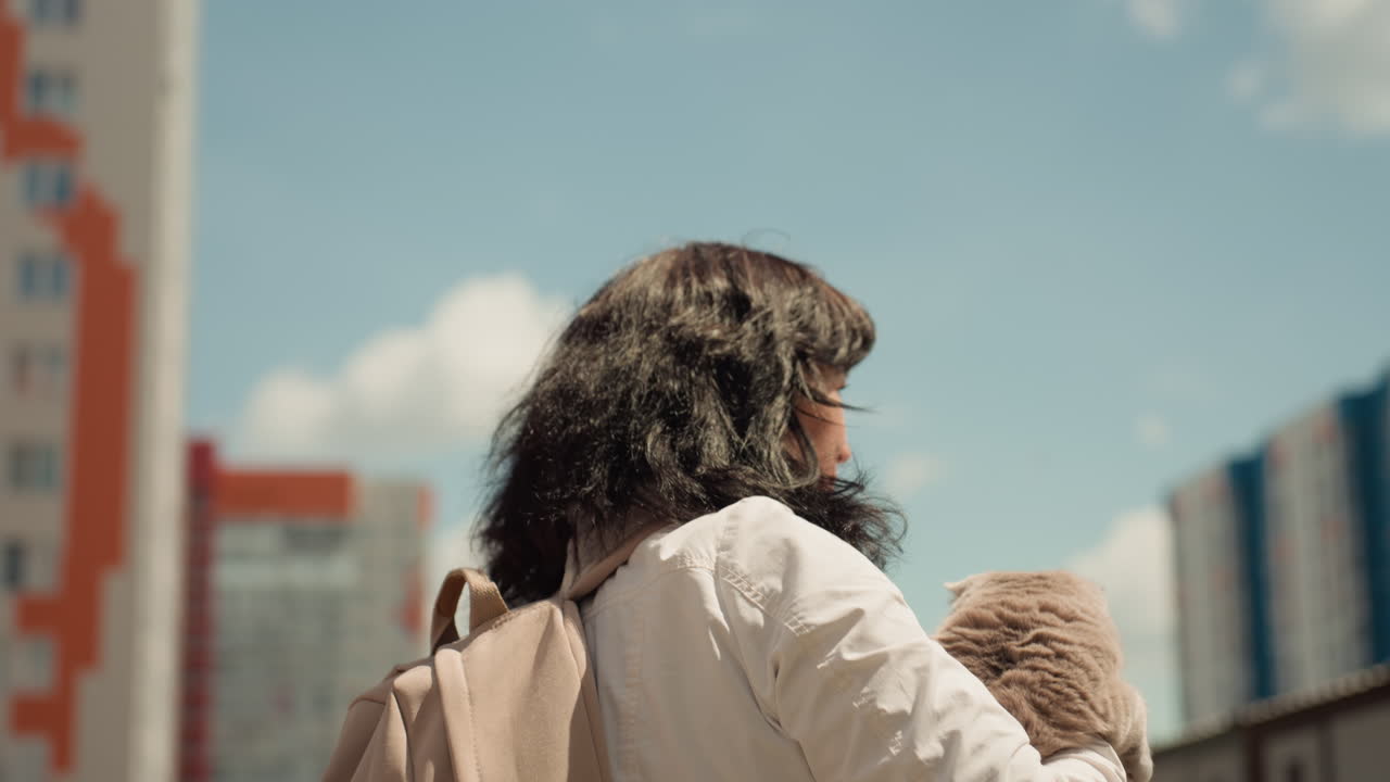 Downtown Scene Featuring Woman And Her Pet, Sunny City Street With Woman Holding Soft Plush Cat And Backpack, Urban Environment Showcasing Woman Walking With Her Cuddly Pet And Travel Bag