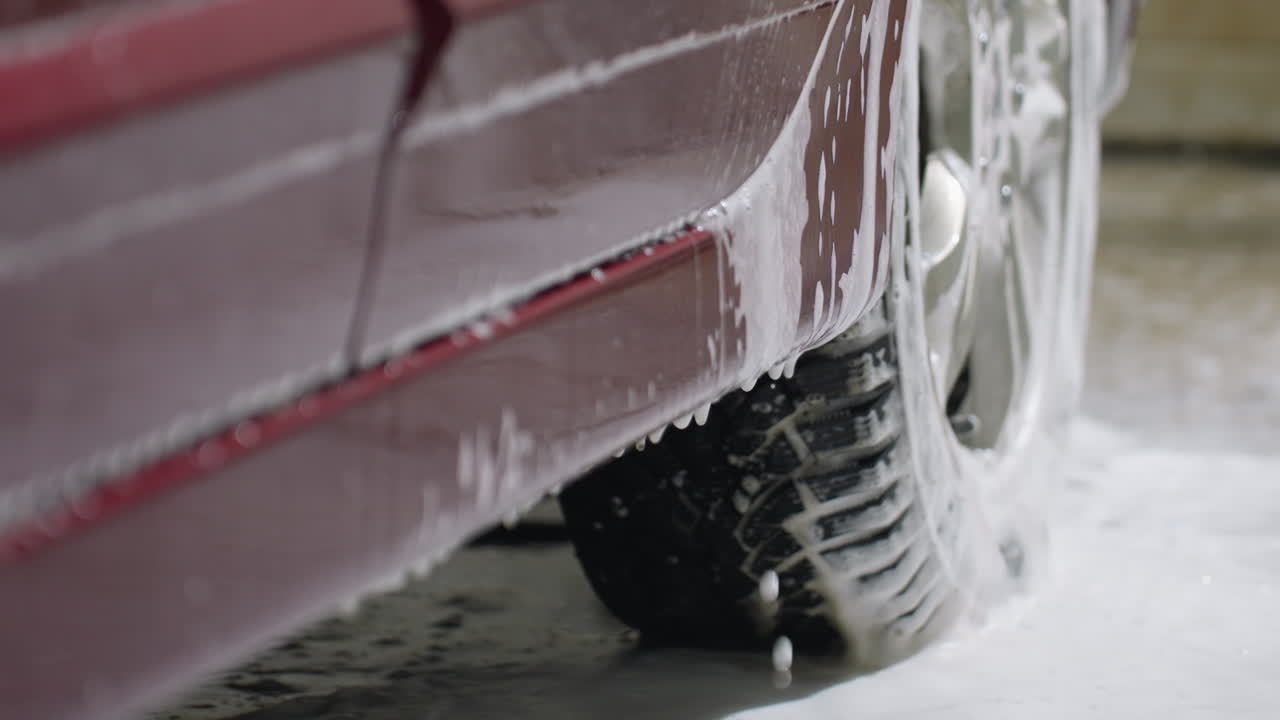 Close up of soapy water dripping from red car body near tire during washing process with suds and droplets creating dynamic texture and fluid flow on vehicle exterior inside garage environment