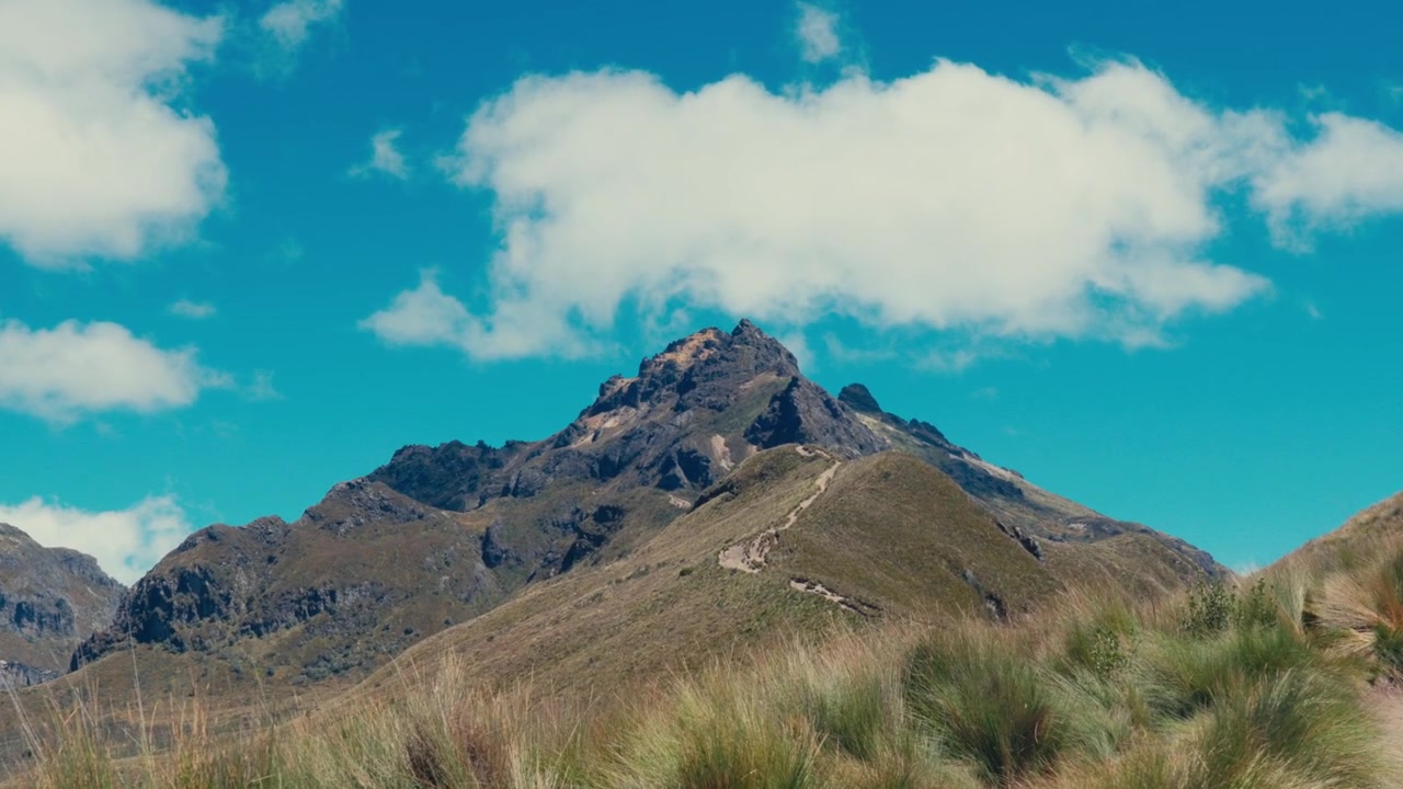 Una vetta montuosa impervia si erge sotto un cielo azzurro brillante con nuvole sparse, vista da un sentiero erboso.