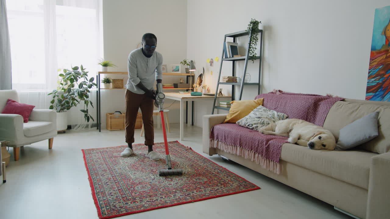 Svart man dammsuger hemma medan hund vilar på soffan