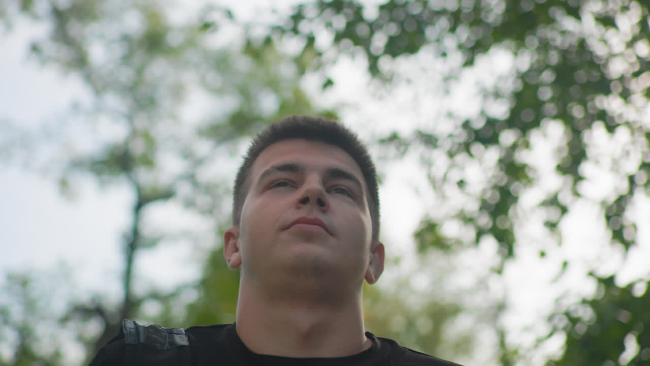 Giovane uomo con zaino che guarda in su attraverso gli alberi; riverbero del sole e bokeh delicato creano una calda retroilluminazione. L'espressione piena di speranza trasmette l'impulso di viaggiare e il processo decisionale in una scena verdeggiante in un parco cittadino.