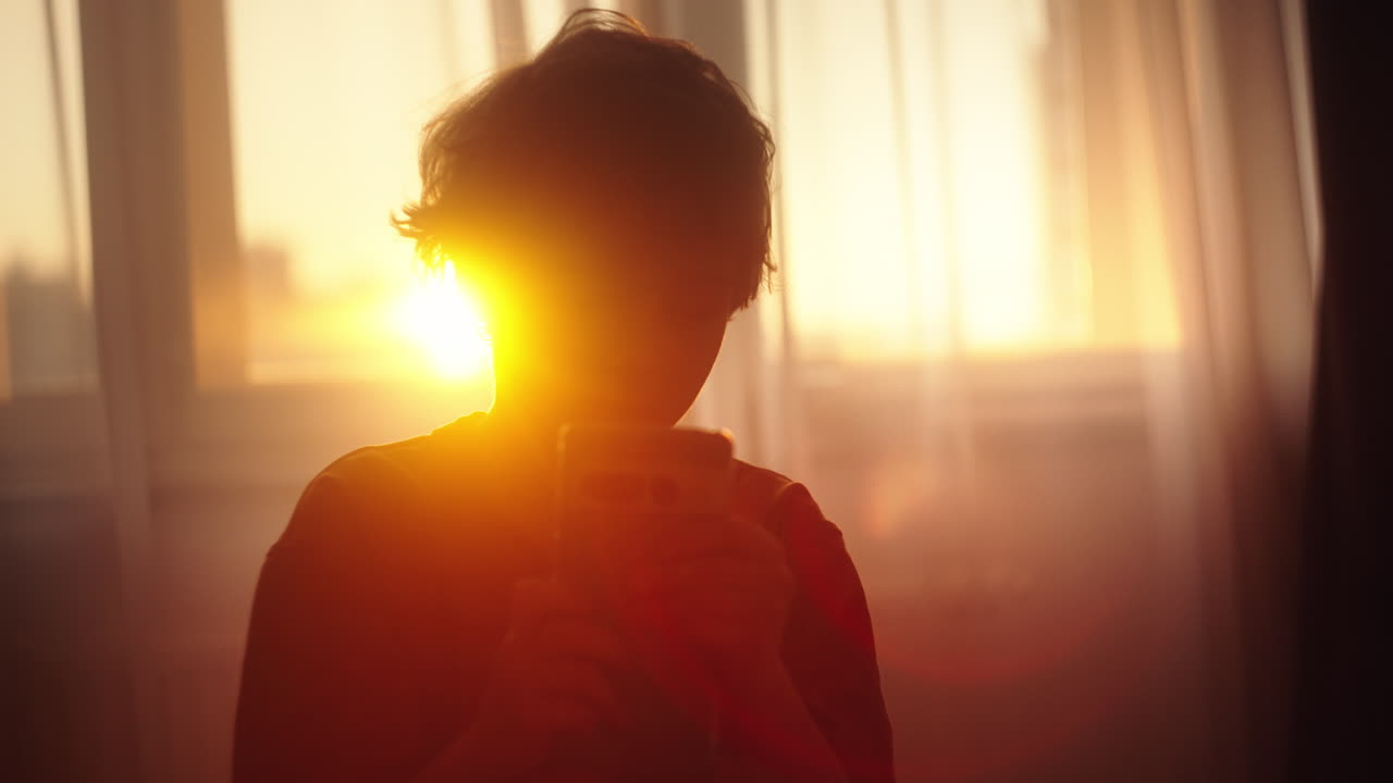 Child Holding Phone at Sunset Through Window