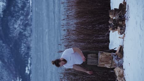 vertical shot of a man stacking chopped woods on snow-covered lake shore in winter