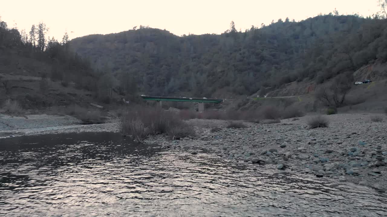 캘리포니아 주 오번에 있는 아메리칸 리버의 녹색 다리를 향해 물과 바위 위를 낮게 날아다니는 드론 - 녹색 나무와 산으로 둘러싸여