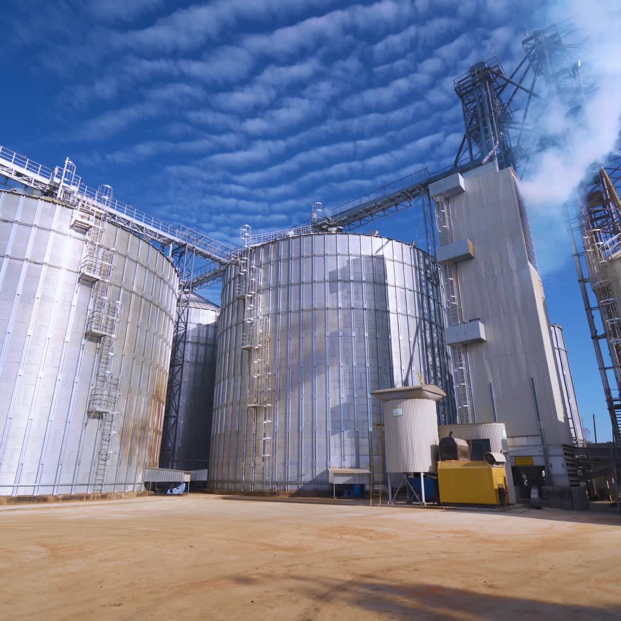 Modern factory under blue sky. Exterior of large grain elevators at sunlight. White vapor goes into the air from industrial plant