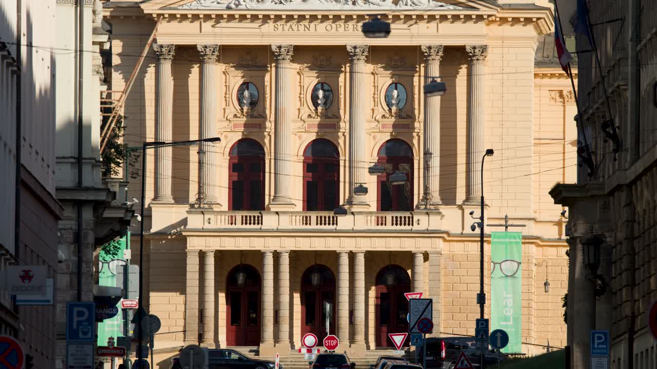 Vehicles drive past neoclassical theater facade in bright daylight, static camera, urban environment