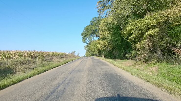 POV (punto di vista) di guida su una strada di contea rurale senza linea centrale che passa accanto a campi maturi, un'aia, alberi e un cimitero nell'Iowa rurale in una soleggiata giornata di inizio autunno