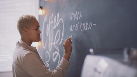 camarera musulmana afro escribiendo tipos de café en la pizarra en un café