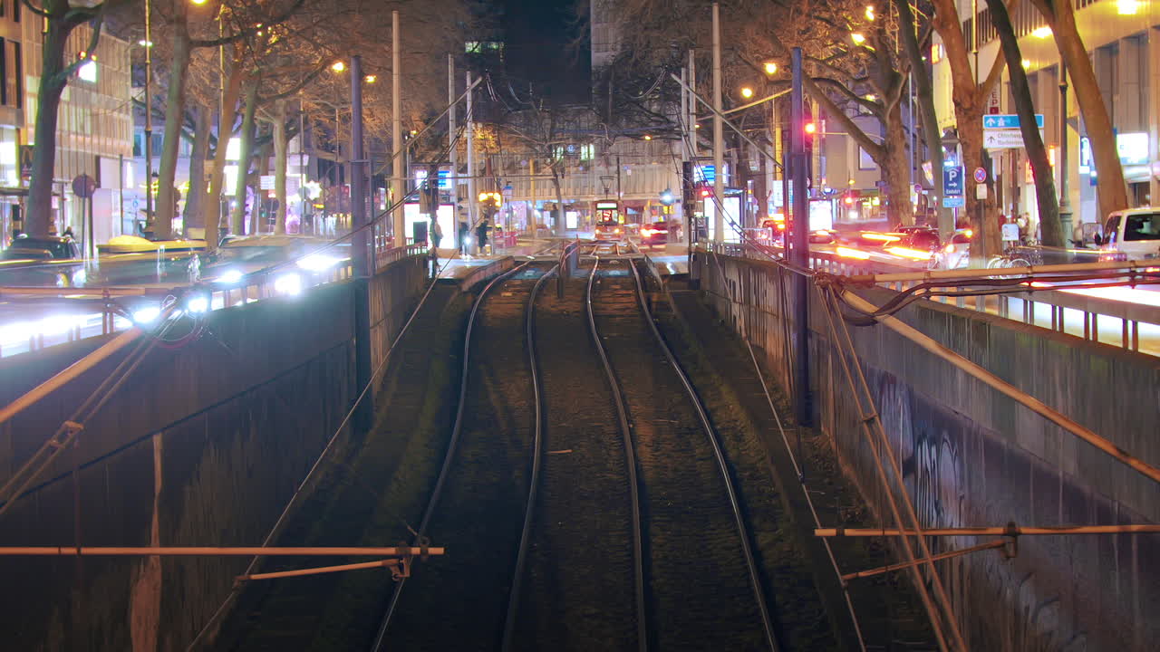 Timelapse with symmetrical arrangement of trams and traffic at the train station. Trams smoothly enter and exit while cars flow on the side. Tracks in the foreground provide a visual focal point. zoom