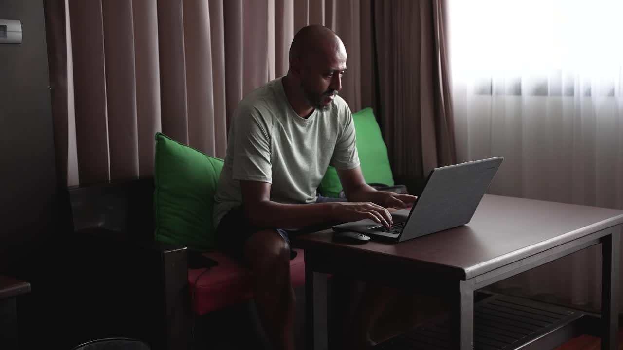 A man comfortably working remotely in a well-lit hotel room, using his laptop while seated at a table