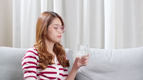 Asian-young-woman-sitting-on-the-sofa-drinking-water-to-relax-during-severe-headache