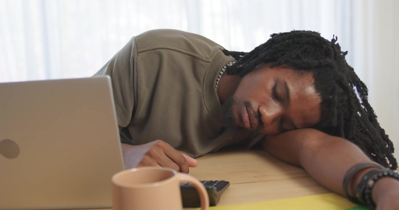 Homme afro-américain reposant sa tête sur son avant-bras à une table après avoir travaillé sur un ordinateur portable, tendant la main vers son téléphone.