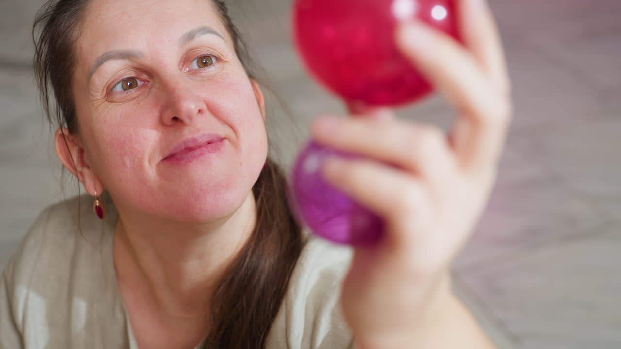 Closeup view of woman smiling warmly while lifting colorful red and purple Christmas ornaments in hand, carefully observing decoration details in cozy home setting