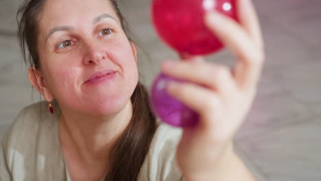 closeup view of woman smiling warmly while lifting colorful red and purple christmas ornaments in hand, carefully observing decoration details in cozy home setting