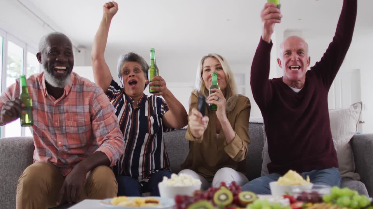 dos parejas mayores diversas sentadas en un sofá viendo un juego bebiendo cerveza