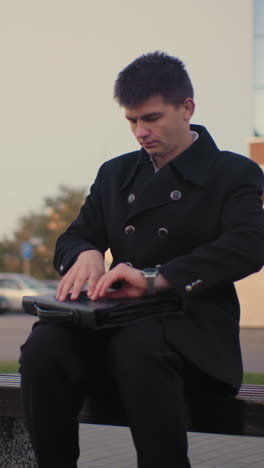 business mogul in black suit walks up to bench, opens briefcase in outdoor setting with modern building and parked cars in background, professional, urban office worker taking a break