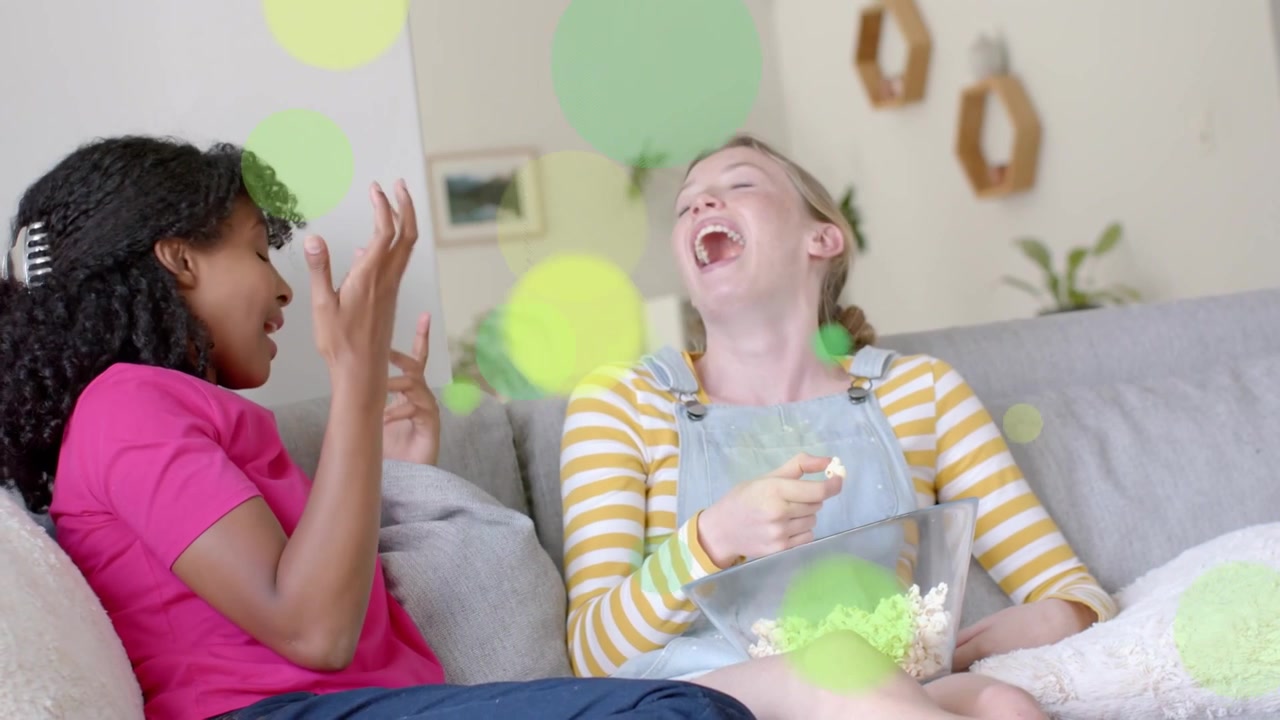 Left girl lifting popcorn to right girl on couch, sharing for leisure as overlays obscuring faces
