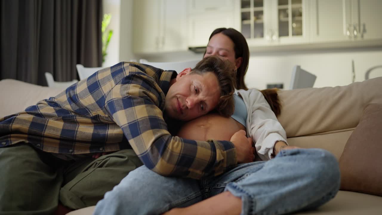 Happy middle-aged man in a plaid shirt listens and leans on the belly of his pregnant wife while communicating with her at home