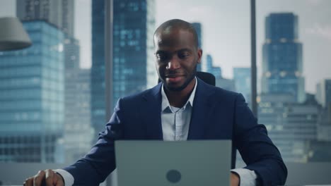 Portrait of Successful Black Businessman Working on Laptop Computer in His Big City Office. Professional Digital Entrepreneur does Data Analysis for e-Commerce Strategy assessment. Zoom In Front View