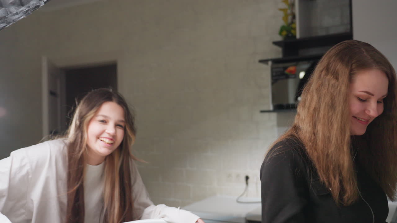 Elegant young lady working in kitchen focused on task as roommate enters with smile giving handshake before climbing chair to adjust ring light, friendly teamwork blending lifestyle activity
