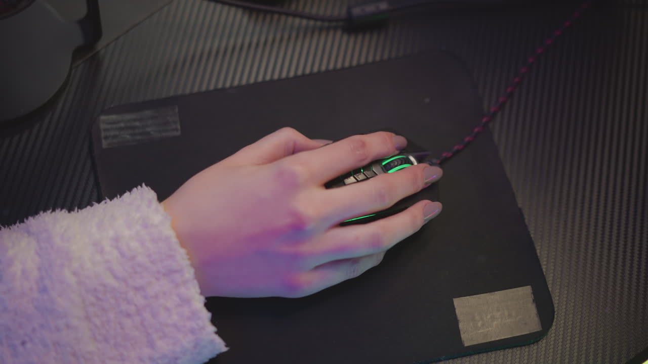 Close up hand of lady in fluffy sweater resting on mouse with green light glowing beneath fingers, placed on black mousepad over dark textured desk, subtle ambient lighting