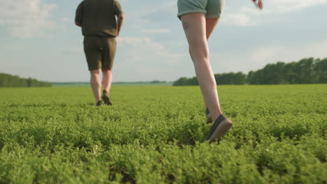 low angle view of friends jogging through lush green field at dusk with woman following behind, legs in motion kicking up grass, emphasizing movement, outdoor exercise and scenic rural landscape