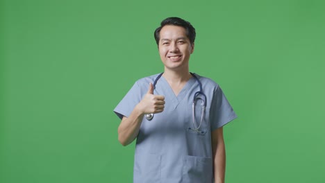 médico masculino asiático con estetoscopio mostrando pulgares hacia arriba gesto y sonriendo en el fondo de la pantalla verde en el hospital