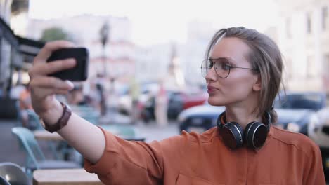 Cheerful-attractive-female-dressed-in-orange-shirt-and-sunglasses-making-selfie-on-smartphone-camera,-posing,-standing-on-street