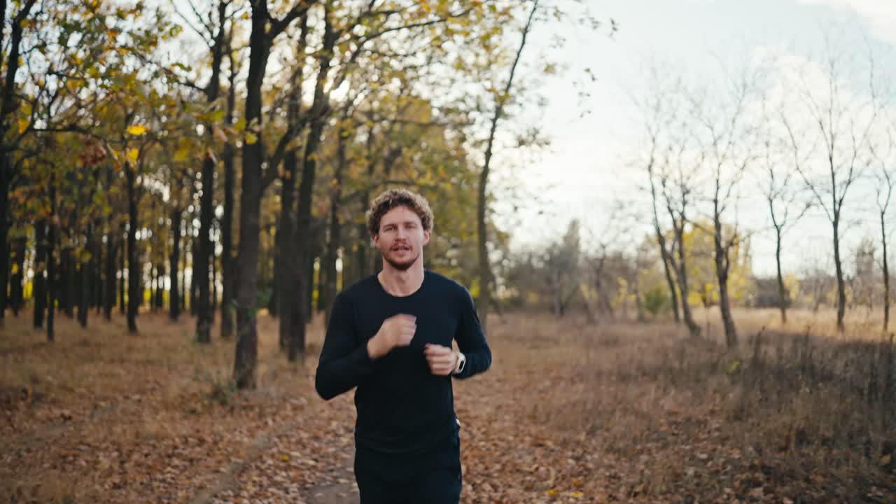 un hombre feliz confiado y ligeramente cansado con cabello rizado y barba en un uniforme deportivo negro mira su reloj durante su carrera matutina en el bosque soleado de otoño