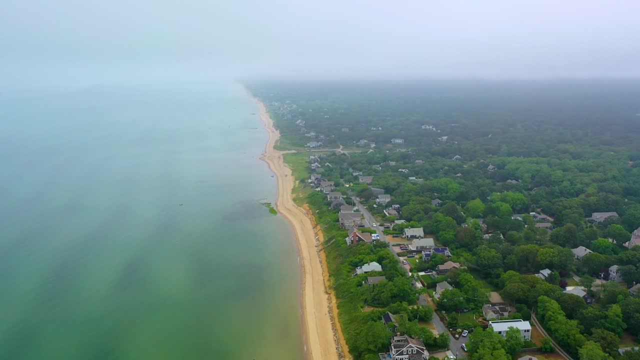 Scenic drone flyover of Cape Cod shoreline under foggy skies, with sandy beach, calm ocean waves, and coastal neighborhoods blending into mist-covered forest and horizon