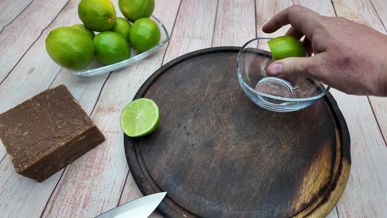 Close-up shot showing human hands slicing a fresh green lime with a knife on a rustic wooden cutting board, next to a block of panela and bowl