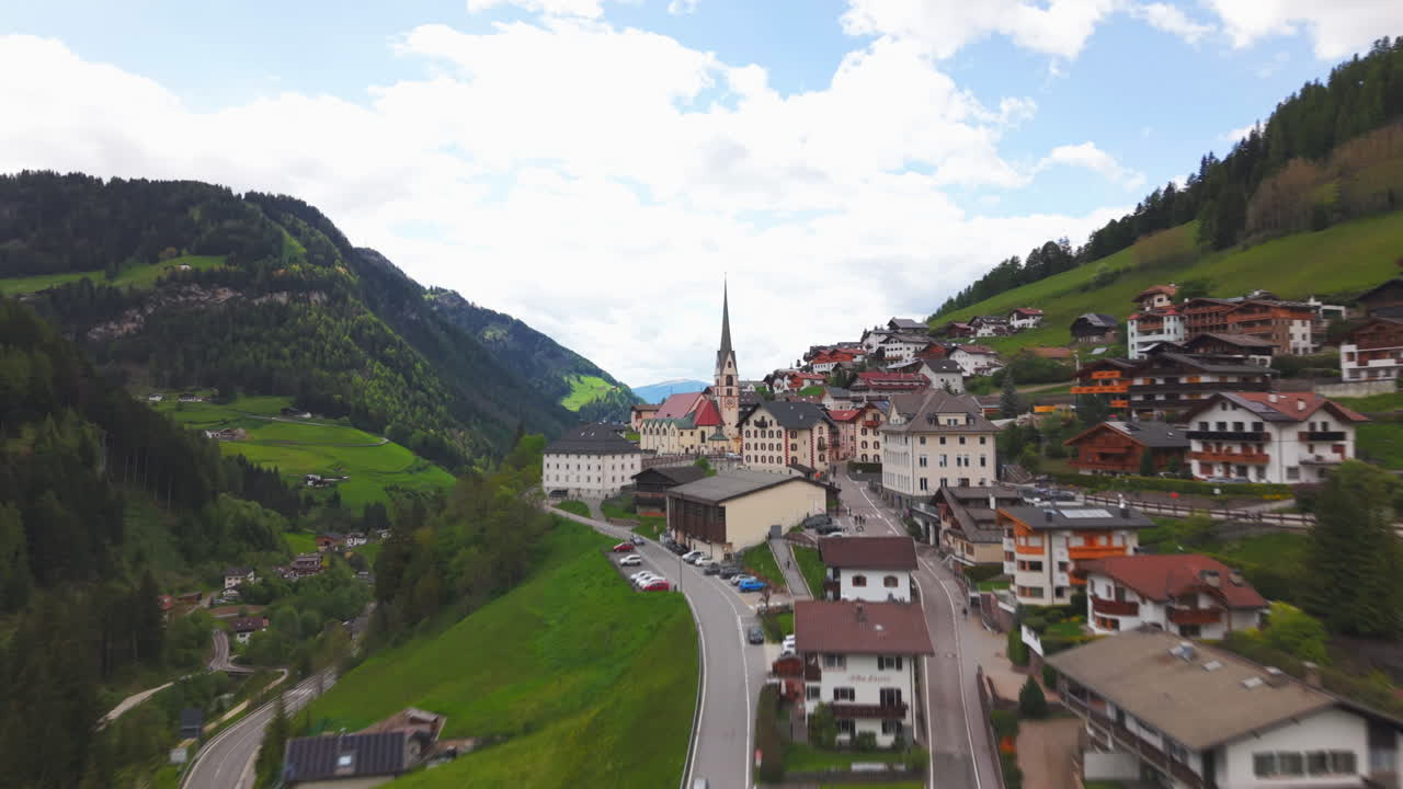 Drone footage of an alpine village with a prominent church and tall steeple, located on a hillside and surrounded by green mountains in the Dolomites, Italy