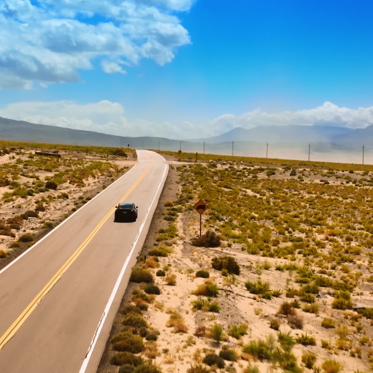 Catching up with a black car driving by the highway in desert. Drone raising above the dry valley in Nevada surrounded by mountains at backdrop