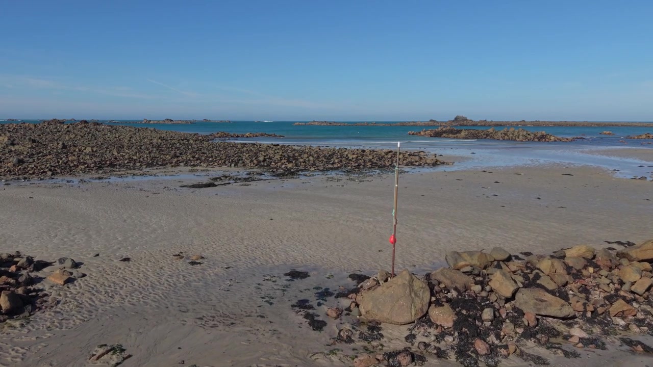Guernsey Kanaløerne. Langsomt fremadgående droneoptagelse ved lavvande, der passerer et sømærke over en sandet forstrand af en barsk klippestrand i sen eftermiddagssol med udsigt over havet og blå himmel.