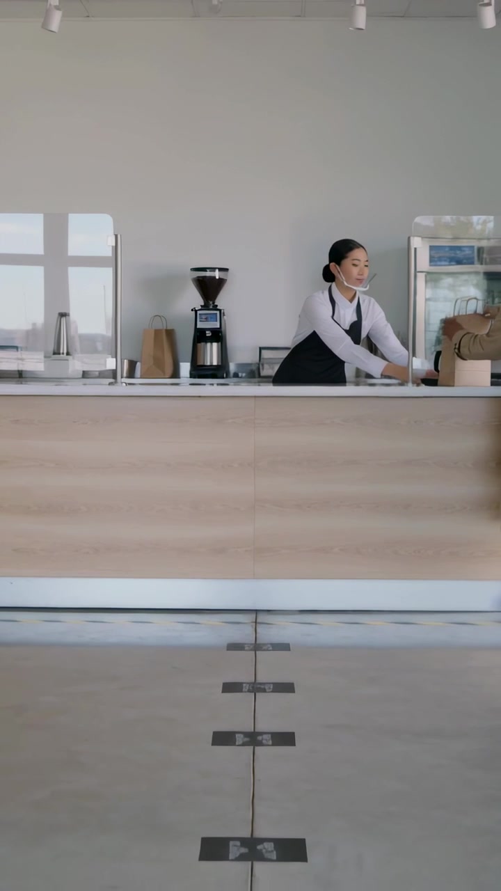 Video vertical: Cliente llegando, barista con delantal sirviendo de una tetera en el mostrador y deslizando una bolsa para el cliente.